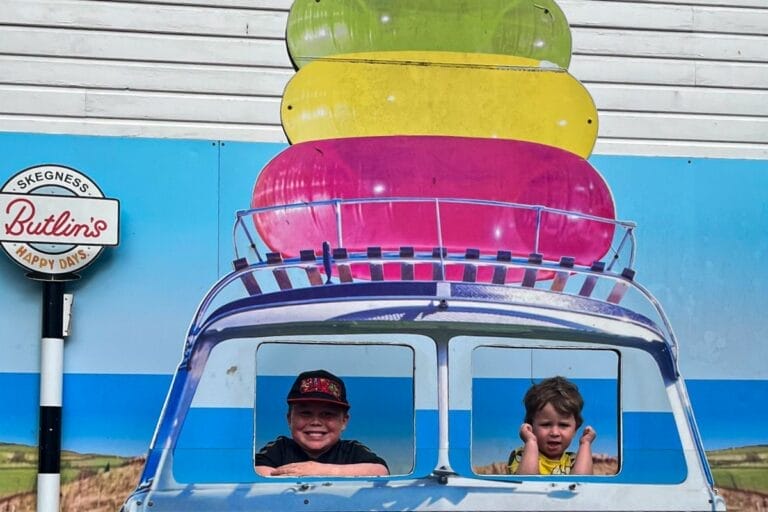 Two children pose behind a cutout of a blue VW van with colorful beach items on top. The backdrop features a scenic landscape. A sign reads "Skegness Butlin's Happy Days." Both kids are smiling and playful.