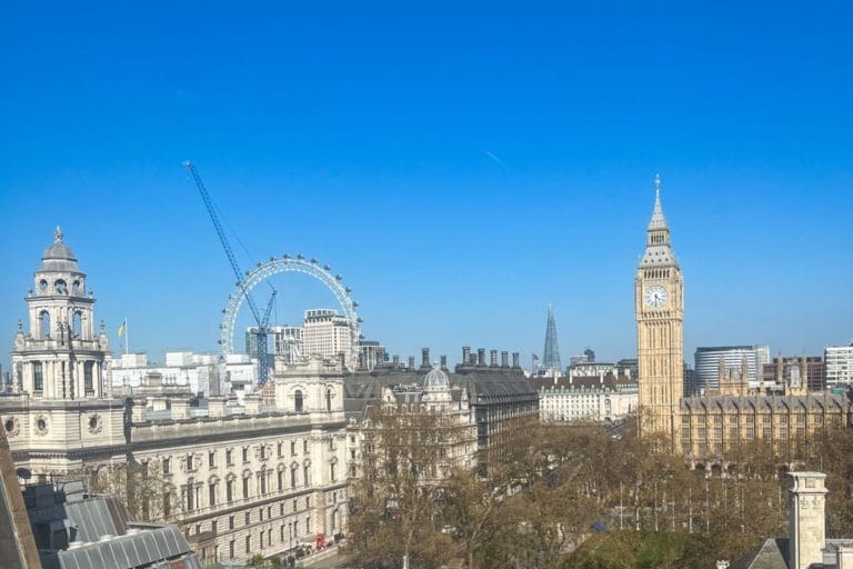 The image shows a clear view of London with the London Eye, Big Ben, and the Shard visible. Historic buildings and trees are in the foreground under a bright blue sky.