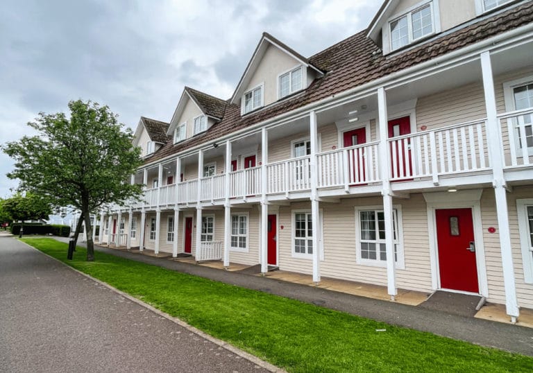 A row of two-story beige apartments with white trim and red doors. Each unit has a balcony and a small porch. A tree and green lawn line the pathway in front of the buildings under a cloudy sky.