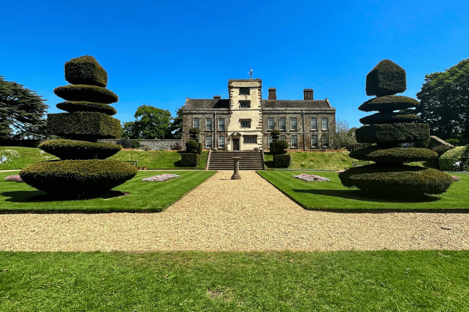 A grand manor house with a neatly manicured lawn, featuring symmetrical topiary trees and a gravel path leading to the impressive entrance, all under a clear blue sky.