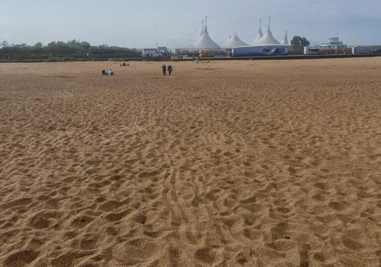 A vast sandy beach with a few people scattered in the distance. Large white tents and buildings are visible at the edge of the beach under a hazy sky. The scene is calm and spacious.