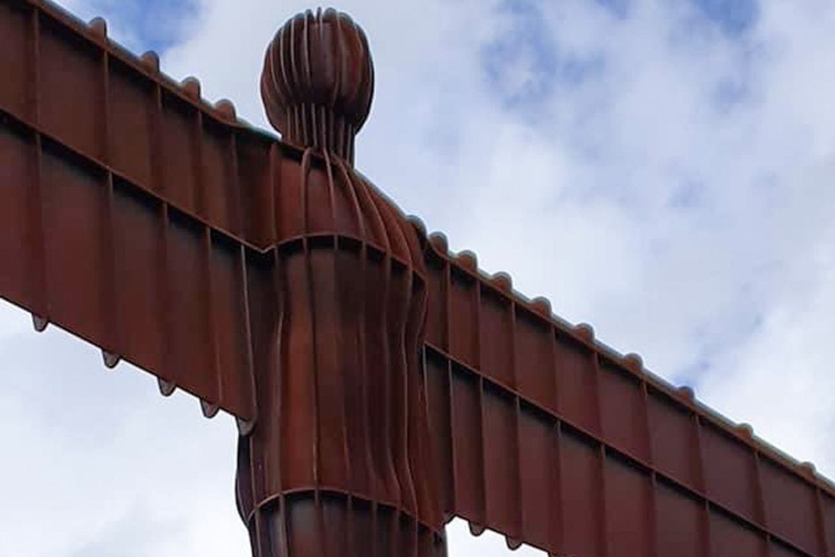 Close-up of the Angel of the North sculpture, a large steel angel with outstretched wings against a partly cloudy sky, showcasing its rust-colored, segmented structure.