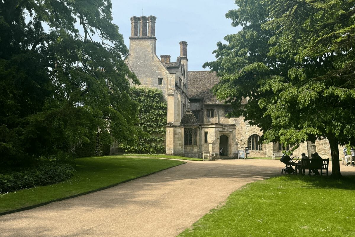 A picturesque view of a historic stone manor house with tall chimneys, partly hidden by large, leafy trees. The house is set within a well-maintained garden with a neatly trimmed lawn. Several people are seated under a tree, enjoying the serene environment on a sunny day.