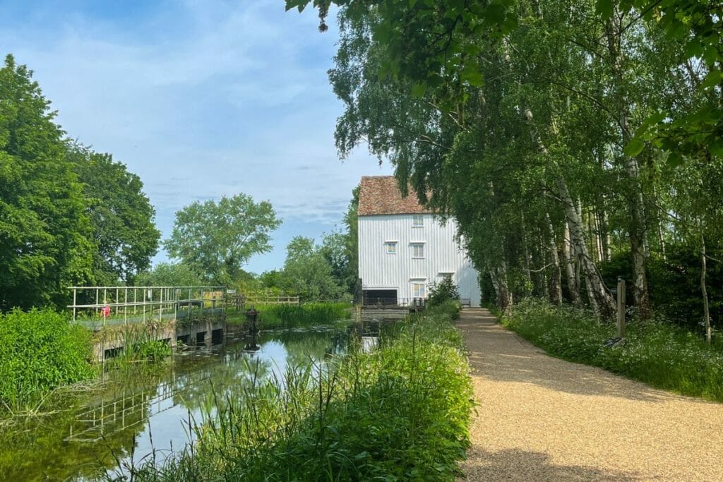 A charming white wooden millhouse standing next to a calm river. The millhouse is surrounded by lush greenery, including tall trees and various plants. A gravel path leads towards the mill, enhancing the peaceful, rural scene.