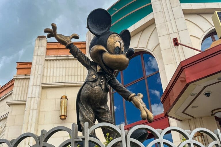 A statue of Mickey Mouse at Disneyland Paris, standing joyfully with arms outstretched in front of an Art Deco-style building with a curved teal roof and large windows reflecting the sky.