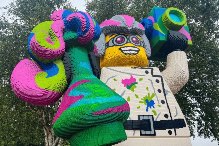 Large LEGO statue of a scientist holding a green and pink beaker in one hand and a blue camera in the other, set against trees.