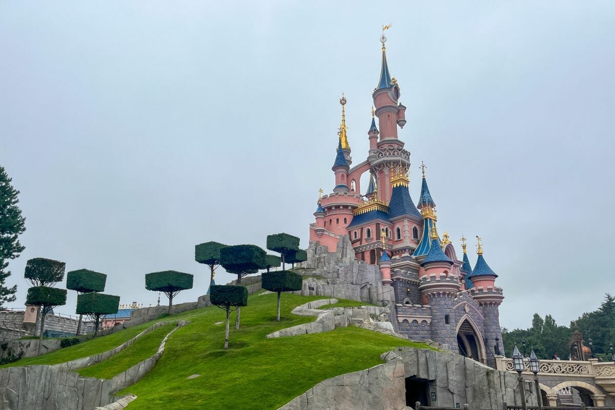 Sleeping Beauty Castle at Disneyland Paris, set on a green hill with manicured gardens and distinctive square-cut trees, under a cloudy sky.