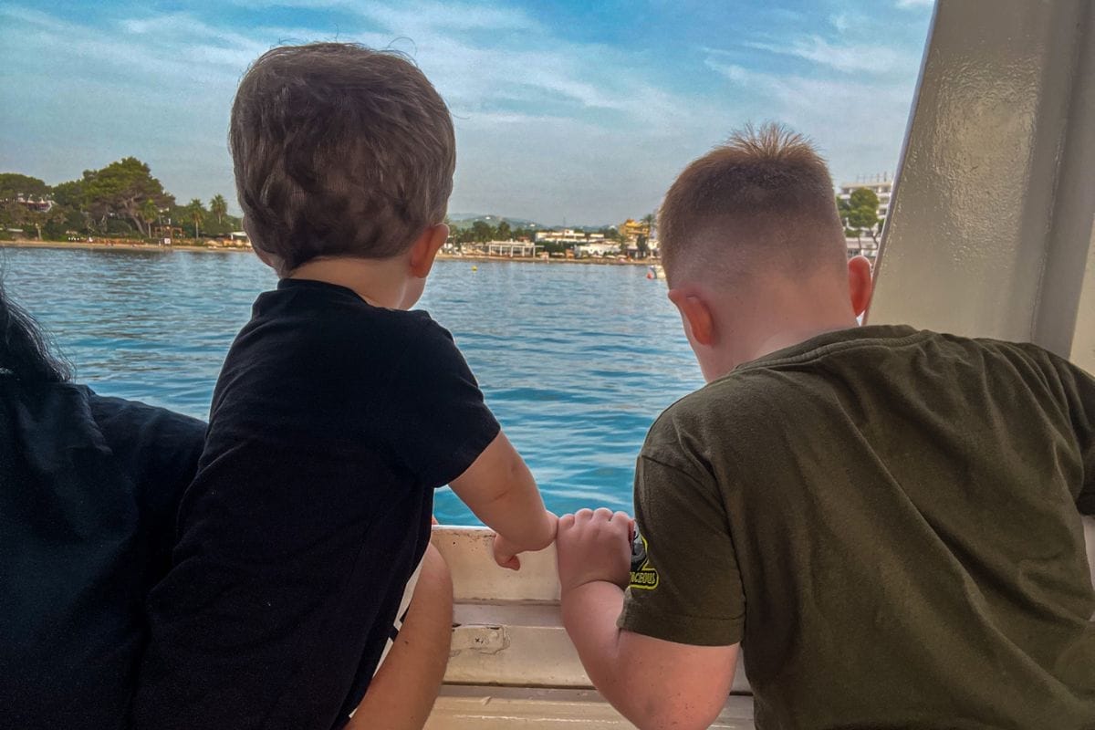 Two children look out over a blue sea from a boat, with one adult partially visible on the left. The coastline with trees and buildings is visible in the distance under a clear, blue sky.