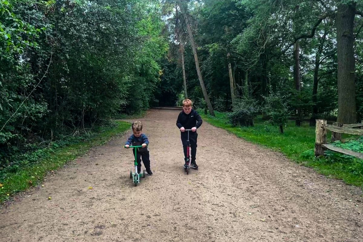 Two children ride scooters on a dirt path in a lush, green forest. The child on the left uses a green scooter, while the child on the right rides a red one. Both children wear dark clothing.