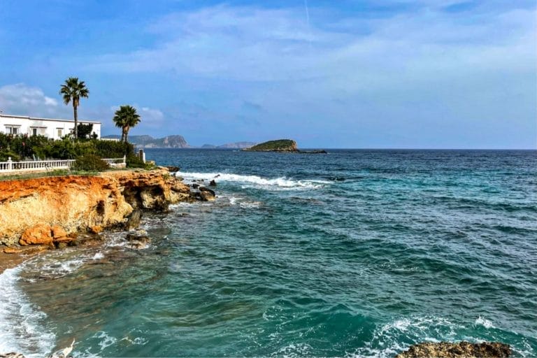 The image shows a rocky coastline of Ibiza with turquoise waves crashing against it. On the left, there are palm trees and a white building with greenery. The sky is clear with distant islands visible.