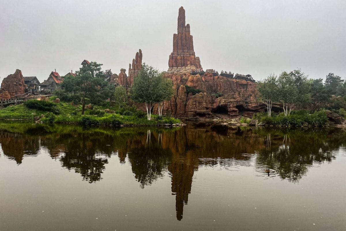 A serene view of Big Thunder Mountain with tall, reddish-brown rock formations reflected in a calm lake. The surroundings are lush with greenery, and the sky is overcast, giving the scene a tranquil and misty atmosphere.