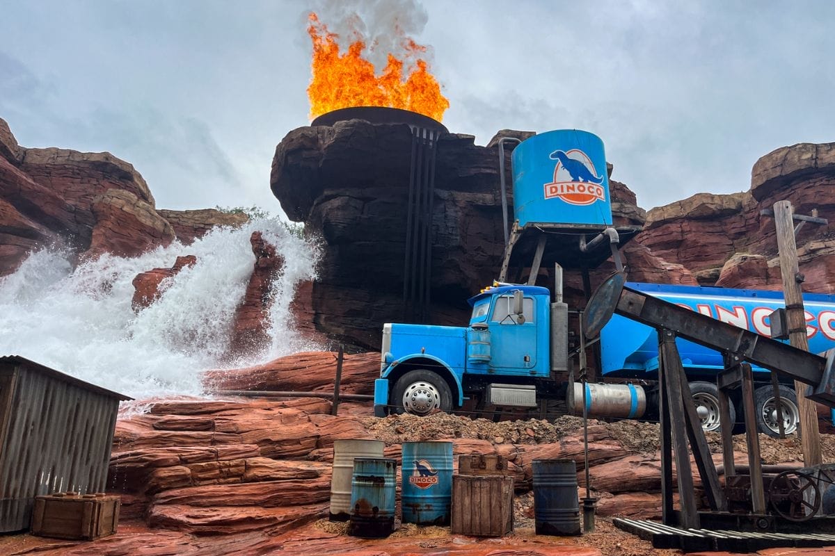 Scene from the Cars Road Trip attraction featuring a large blue truck with "Dinoco" branding, fake fire, and water effects in a rocky desert setting.