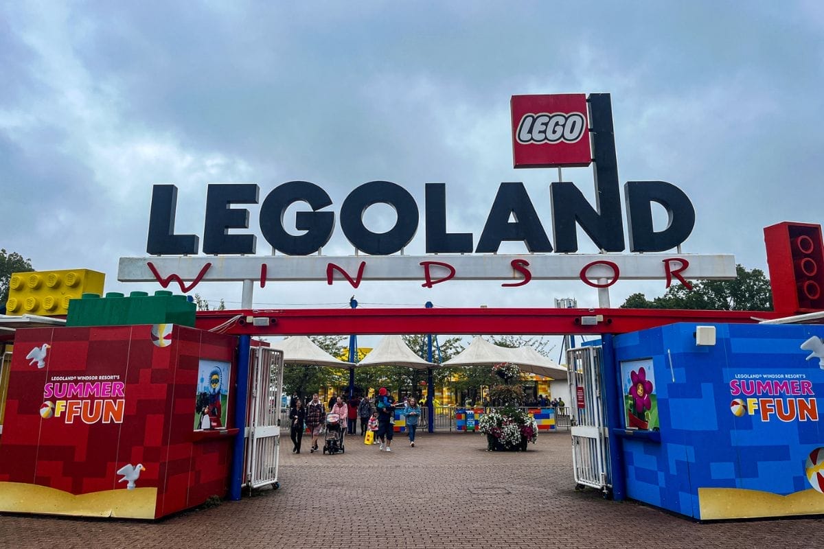 The main entrance to Legoland Windsor with a large sign and colorful Lego bricks. People are entering under cloudy skies, and a booth promoting "Summer of Fun" is visible.