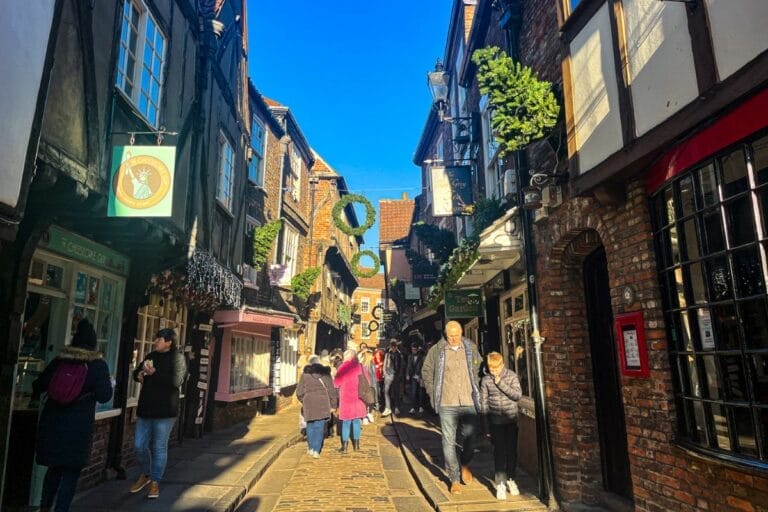 A bustling narrow street with old brick buildings, filled with people walking and shopping. The street is adorned with festive decorations, and the sun is shining brightly.