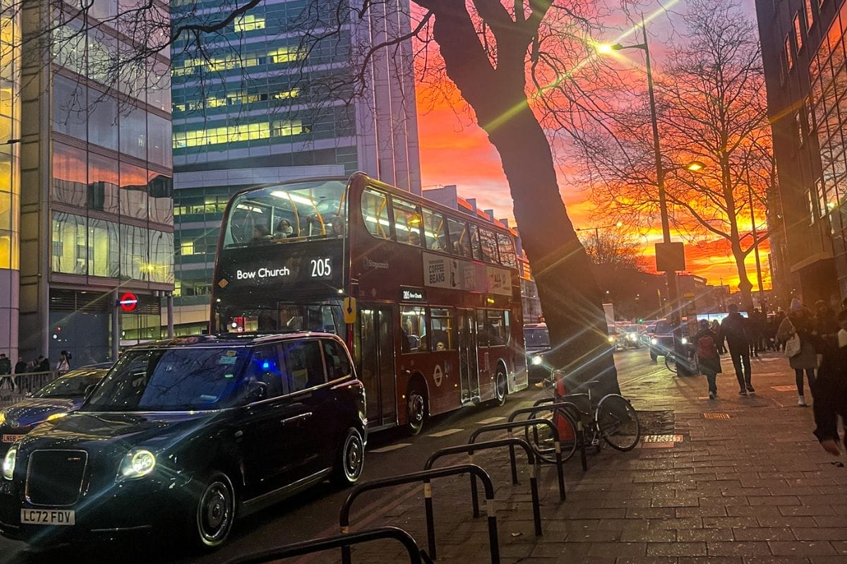 A vibrant sunset over a London street with a red double-decker bus and a black taxi. The sky is ablaze with pink, purple, and orange hues, casting a glow over the cityscape and tall buildings.