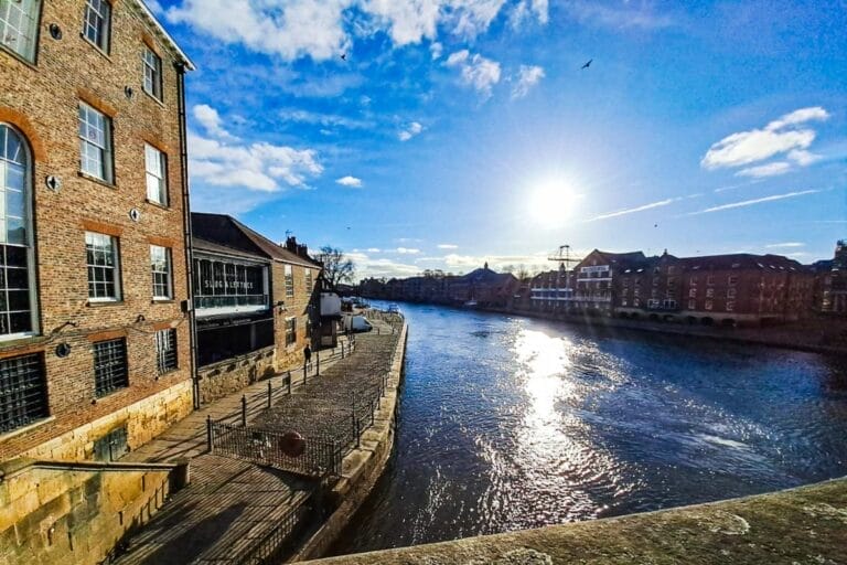 A view of a river with historic buildings on both sides under a bright blue sky. The sun is low, casting a warm glow on the water, and a few birds are seen in the sky.