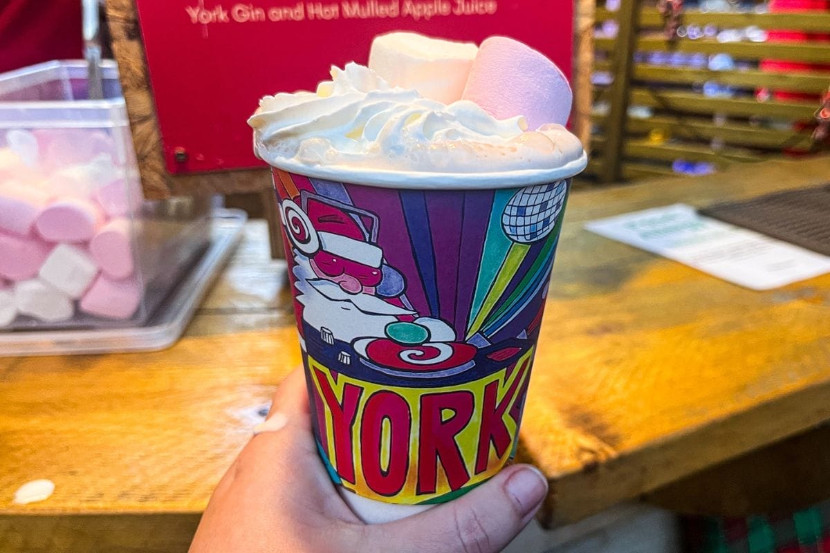 A colorful festive drink in a cup labeled "YORK," topped with whipped cream and marshmallows, held by a hand. The background includes a wooden counter and a sign.