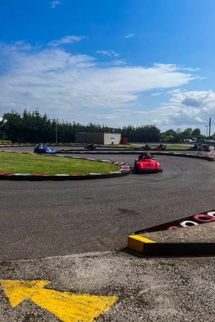 Outdoor go-kart track with several karts racing around the curves. A red go-kart labeled "4" is in the foreground, with blue and black karts behind. The track is bordered by red and white barriers under a clear blue sky.