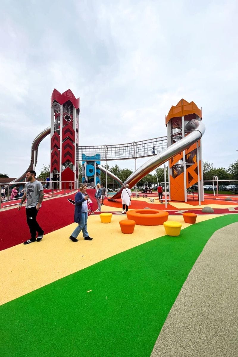 Colourful playground at the Sky Park at Butlins. There are tall red and orange towers connected by a bridge and large slides. People walk and play on the vibrant ground, featuring various shapes and play structures under a cloudy sky.