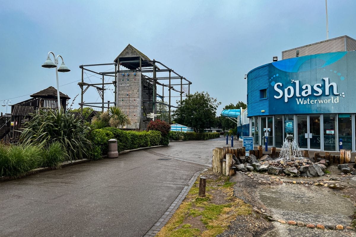 A wide-angle view of the entrance to Splash Waterworld at Butlins Minehead. A building with a blue façade and the Splash Waterworld sign stands prominently on the right, while an outdoor climbing structure and lush greenery line the path on the left.