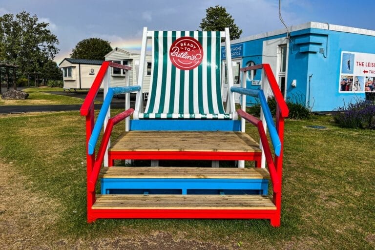 A giant colorful deck chair in Butlins Minehead. The chair has red, white, and blue stripes, and a logo in the center with the words "Ready for Butlins." Surrounding the chair are small cabins and a bright blue sky.