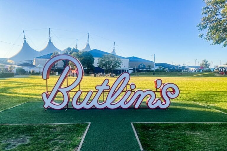 The iconic Butlins sign at Minehead, set on a lush green lawn. The sign is large, with bold red letters outlined in white, positioned in front of the resort's white tent-like structures.