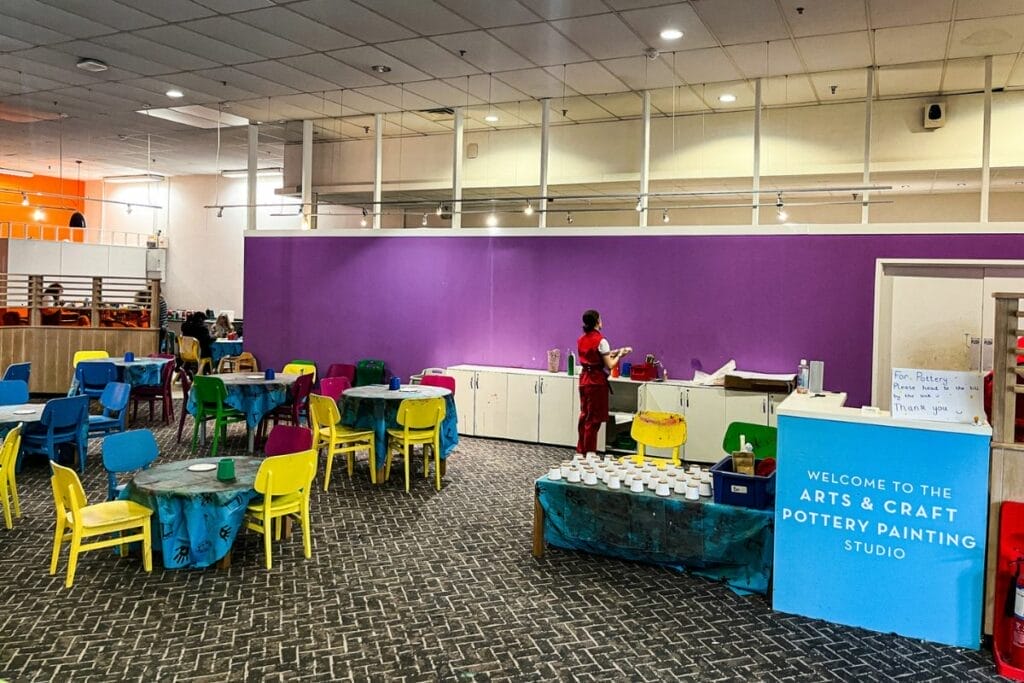 An arts and crafts studio at Butlins Minehead. The room features colorful chairs, tables with protective cloths, and a purple accent wall. A staff member stands by a counter labeled "Arts & Craft Pottery Painting Studio."