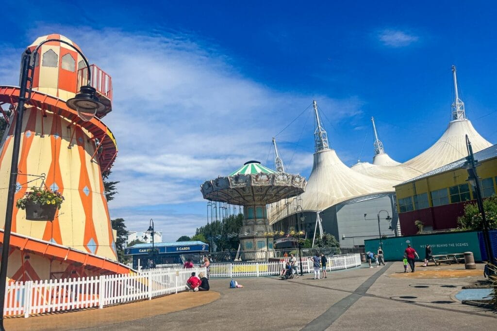 A vibrant fairground scene at Butlins Minehead featuring a classic red-and-yellow-striped helter-skelter slide on the left and a vintage swing ride in the center. The white tent structures are visible in the background under a blue sky.