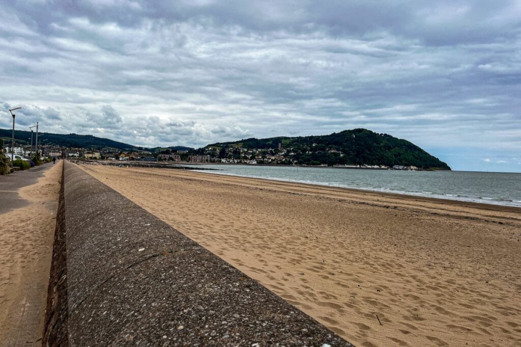 A panoramic view of Minehead Beach, showcasing a wide expanse of golden sand leading to the ocean. A concrete seawall runs parallel to the beach, with lush green hills in the distance under a cloudy sky.