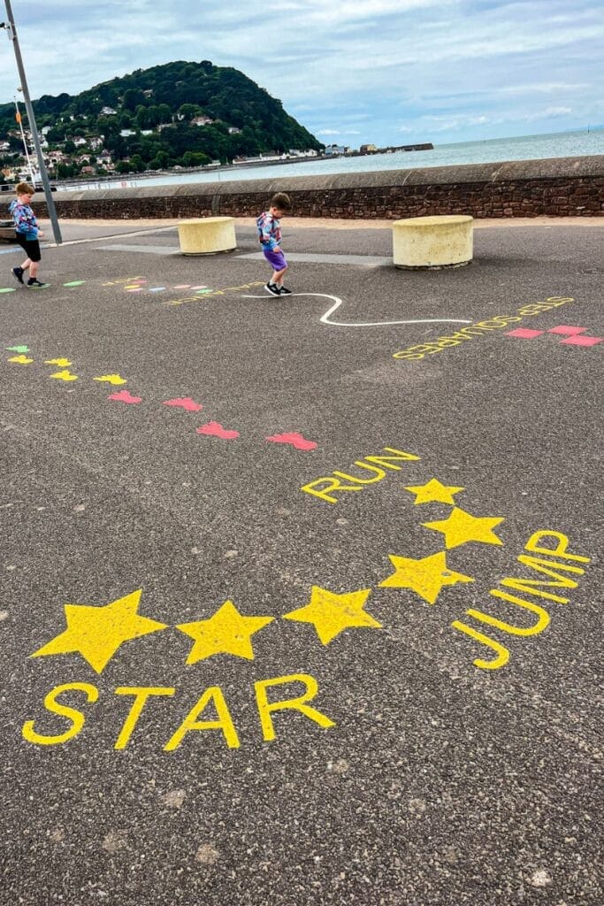 A scenic pathway along Minehead Beach, featuring colorful markings for a children's play area labeled "Star Jump." Two children wearing matching tops play near the seawall, with a hilly landscape and ocean in the background.