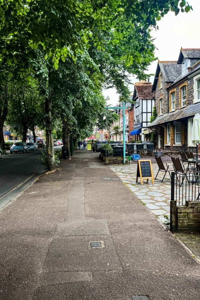 A quaint street scene in a town center features a wide sidewalk lined with trees on the left and a row of charming old buildings on the right. Outdoor seating with chairs and tables is visible, along with a chalkboard sign advertising a menu. The street has light traffic and a few pedestrians in the distance, with a canopy of green leaves overhead providing shade.