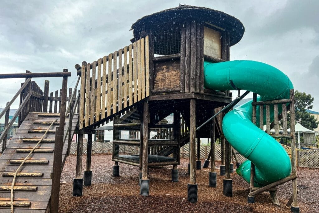 A wooden play structure at Butlins Minehead with a green spiral slide and a series of platforms connected by bridges and ladders. The area is surrounded by wood chip mulch, providing a safe play surface for children.