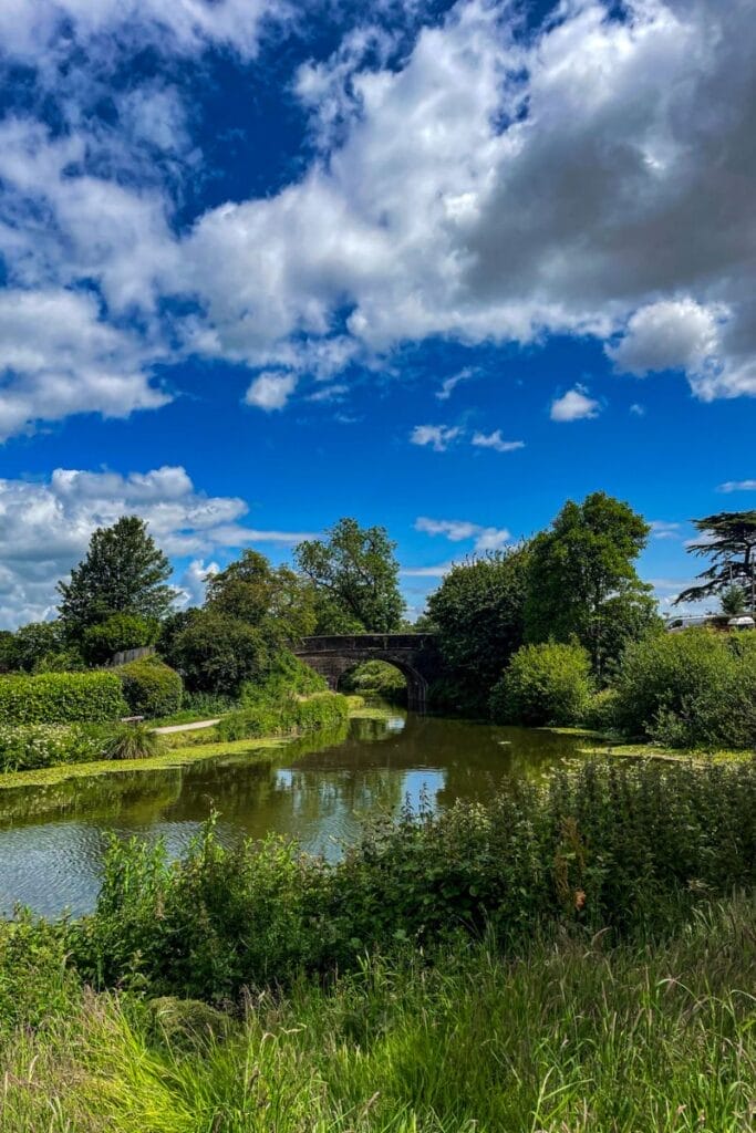 The image shows a tranquil countryside scene with a stone bridge arching over a calm river. The river is surrounded by lush green vegetation, and the sky above is a vibrant blue with fluffy white clouds scattered across. Tall trees line the riverbank, adding to the serene, natural setting.