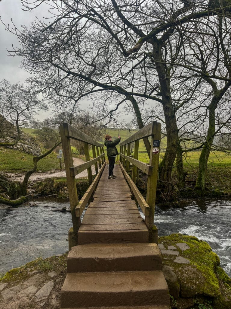 A person in a green jacket stands on a wooden footbridge over a flowing stream, taking a photo. Leafless tree branches frame the scene, with grassy hills and a cloudy sky in the background.